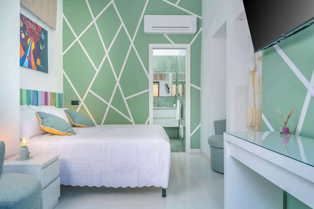 A modern bedroom with a geometric wall design, a bed with pillows, a bedside table with a lamp, and a bathroom visible through an open door.