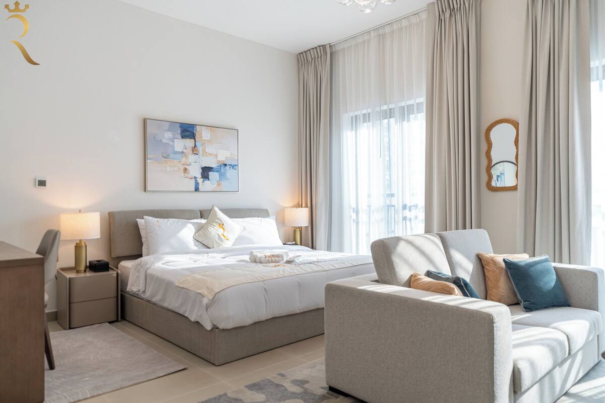 A modern hotel room with a bed, two bedside tables, a sofa, a large window with curtains, and a wall-mounted mirror.