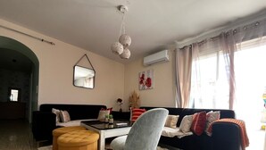 Ett vardagsrum med en soffa, en pall, en fåtölj och ett soffbord. På väggen hänger en spegel, det finns en luftkonditioneringsanläggning och ett fönster med gardiner.
