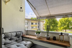 Un balcon doté d’un espace salon avec des coussins, d’une plante en pot, d’une lanterne et offrant une vue sur un immeuble avec des balcons.