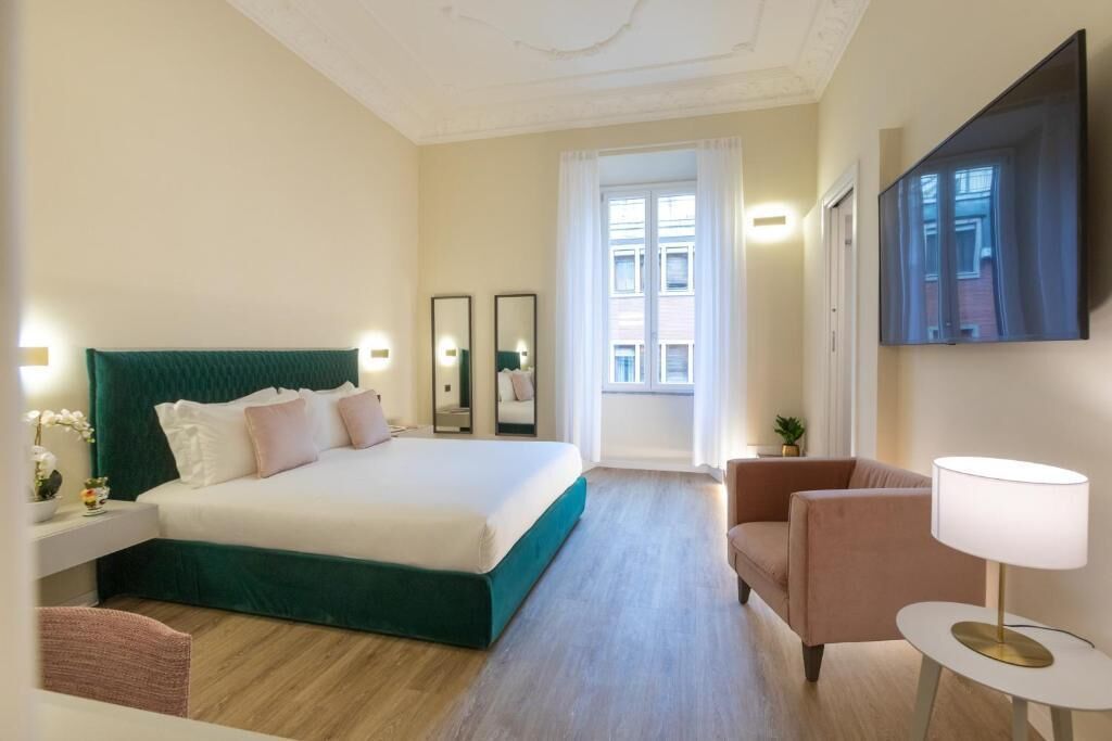 A hotel room with a large bed, a sitting area, a TV mounted on the wall, and a window with curtains.