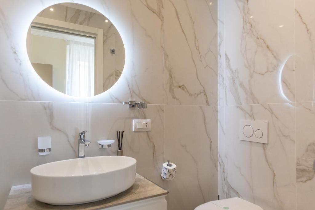 A bathroom with a round mirror, a white sink, a toilet, and a marble wall.