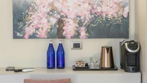 Un tableau représentant des fleurs de cerisier, accroché au mur, une étagère blanche avec une machine à café, deux bouteilles bleues et une chaise.