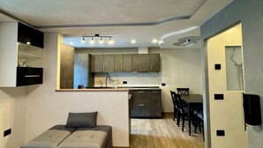 A modern living room with a sofa, a dining area with chairs, a kitchen with cabinets, and a ceiling light fixture. - Apartment "Cima Piazzi" – Comfort, Relaxation, and Nature between Bormio and Livigno (Semogo)