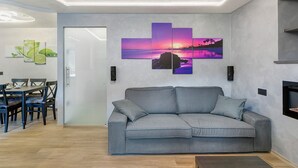 A modern living room with a grey sofa, a dining area with a table and chairs, and a wall with a sunset painting. - Apartment "Cima Piazzi" – Comfort, Relaxation, and Nature between Bormio and Livigno (Semogo)