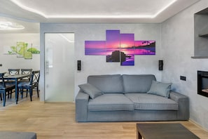 En moderne stue med en grå sofa, et spiseområde med bord og stole, og en væg med et solnedgangsmotiv.