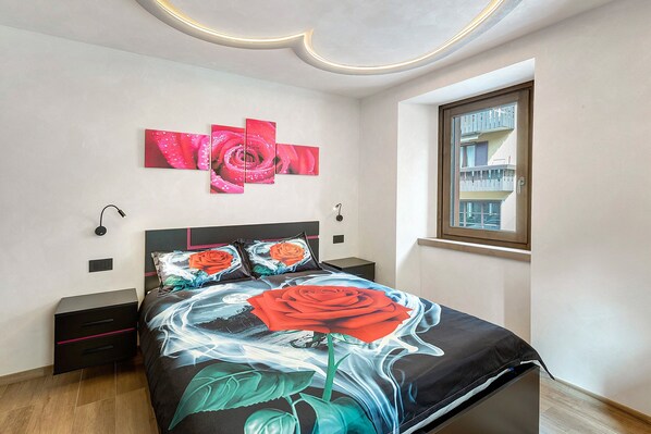 Quarto com cama, mesa de cabeceira, obra de arte na parede, janela e vista para varanda.