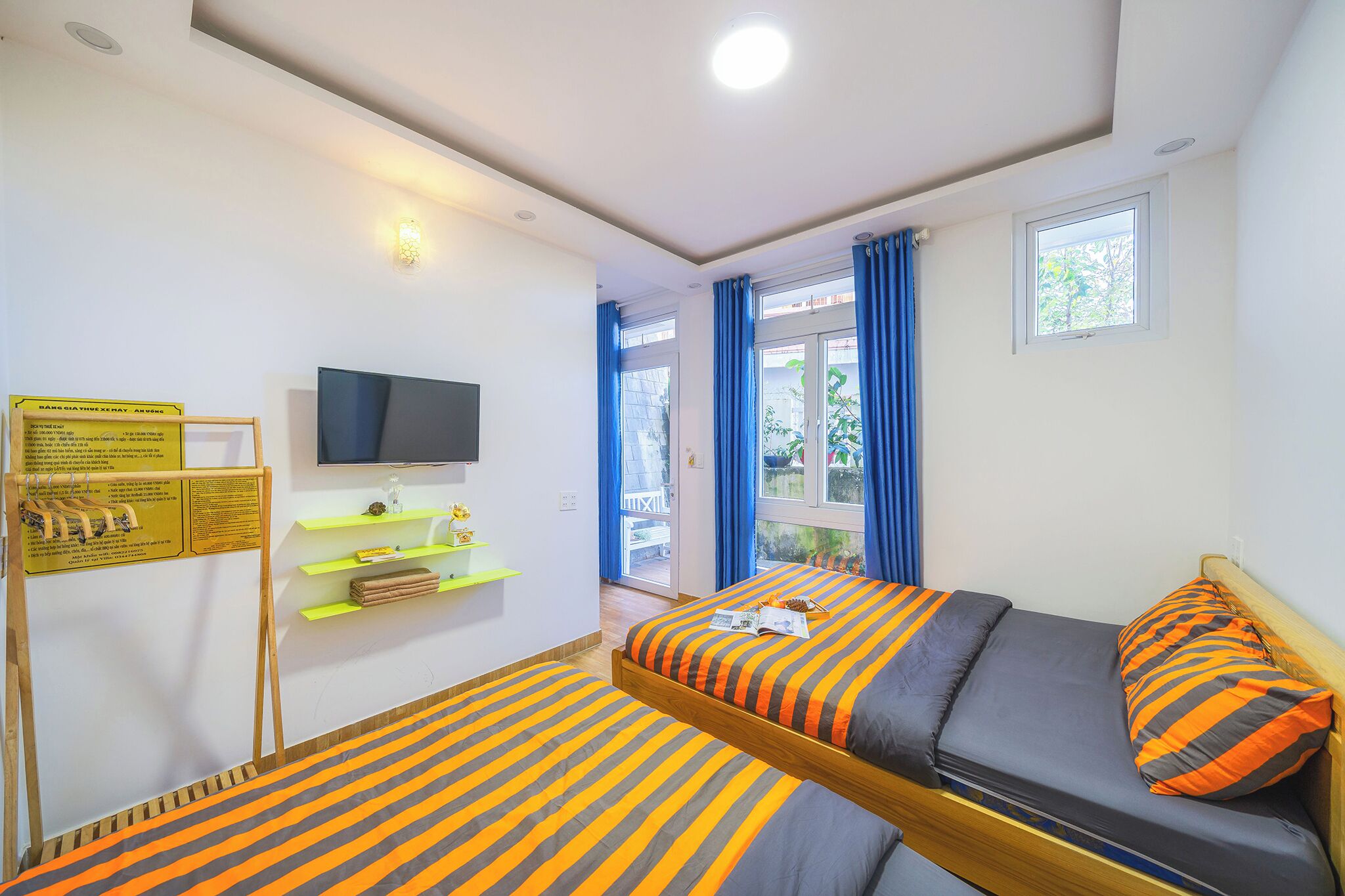 Una habitación con una cama, televisión, ventana con cortinas, estante con objetos y un cartel en la pared.