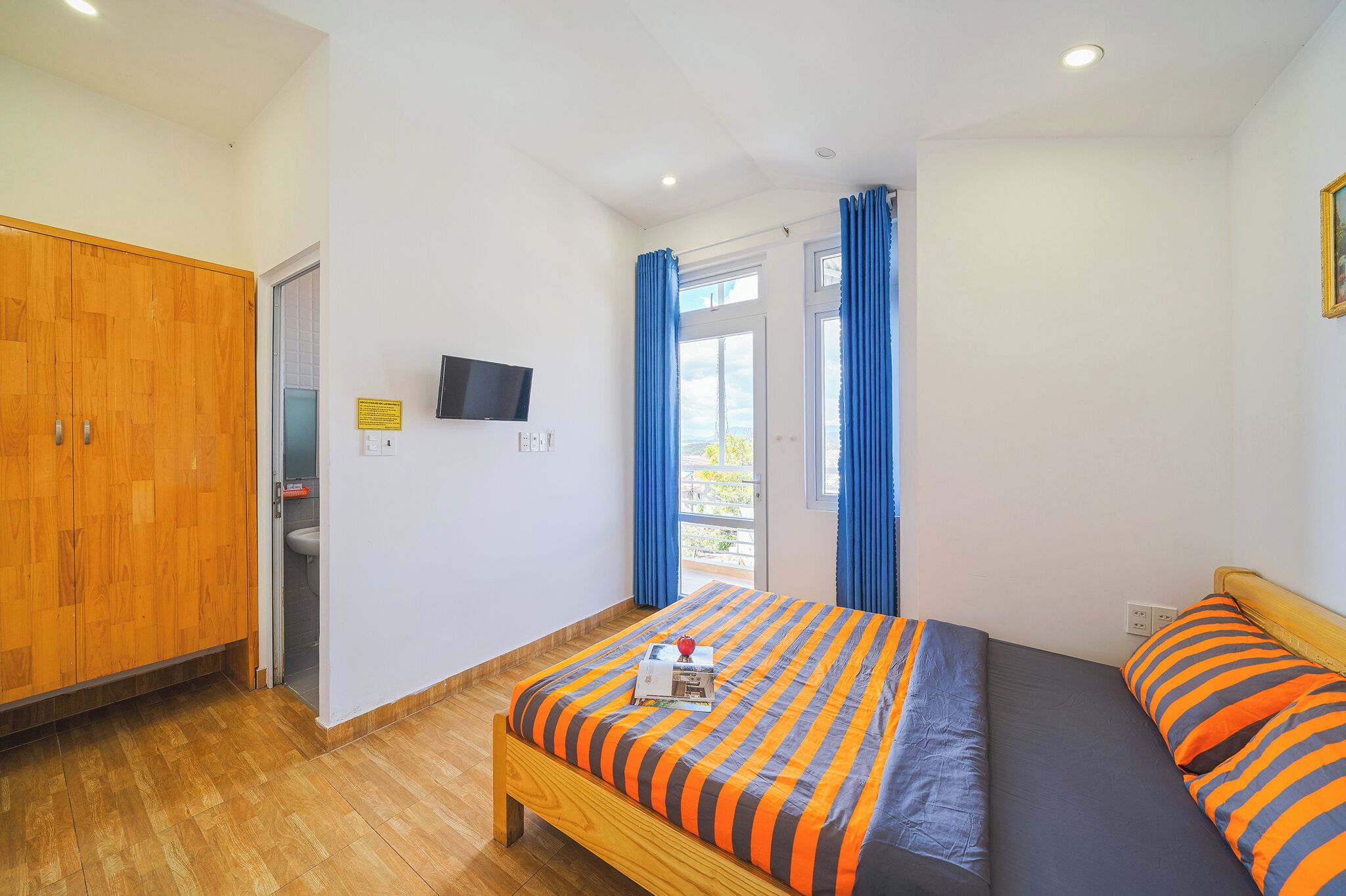 A bedroom with a wooden wardrobe, a bed with striped bedding, a television on the wall, and a window with blue curtains.