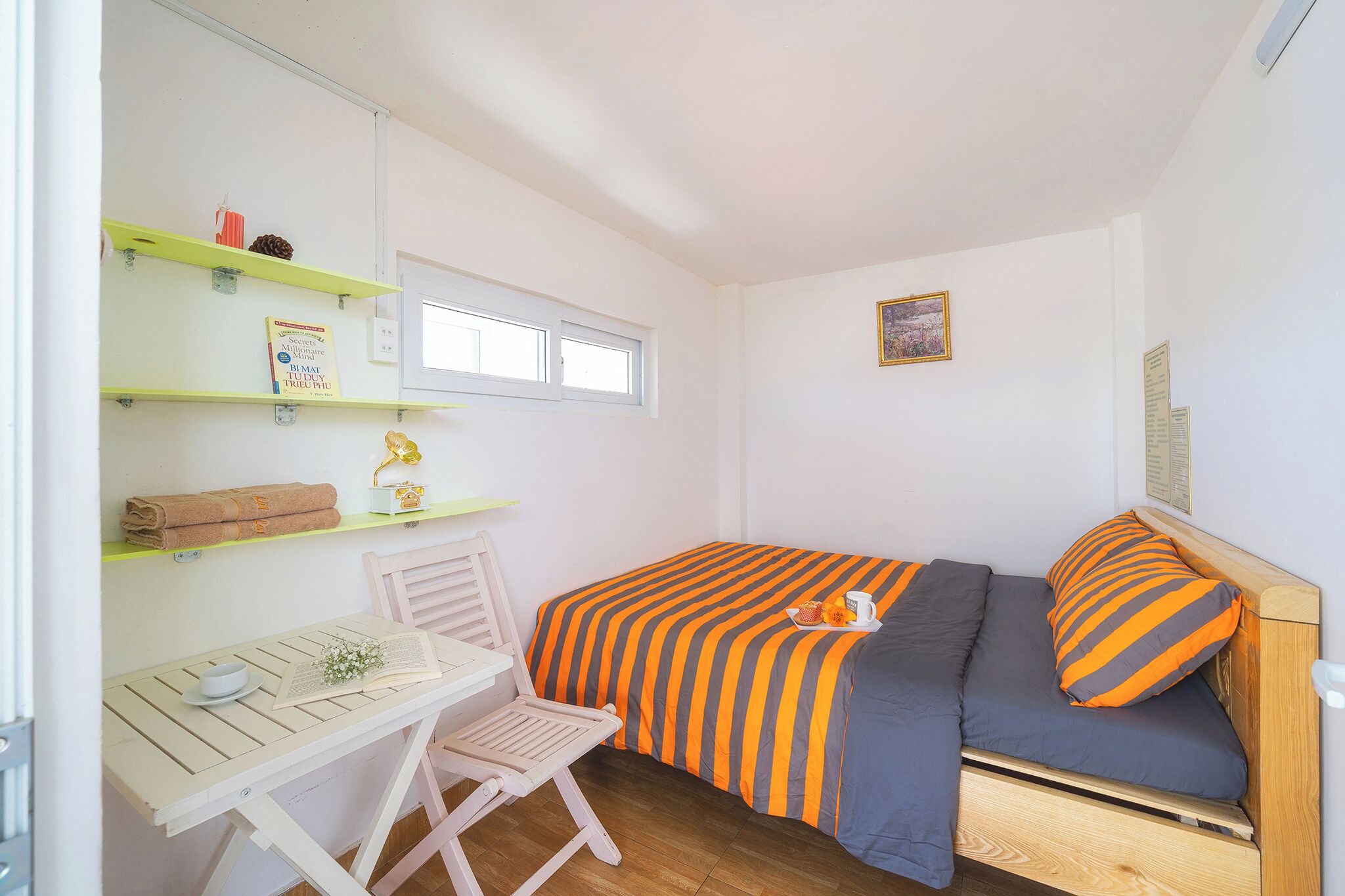 Een kleine, verzorgde kamer met een bed, een bureau en een stoel. Er staan planken met spullen op, een raam en een ingelijst schilderij aan de muur.