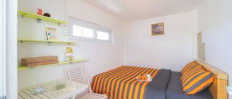 Een kleine, verzorgde kamer met een bed, een bureau en een stoel. Er staan planken met spullen op, een raam en een ingelijst schilderij aan de muur.