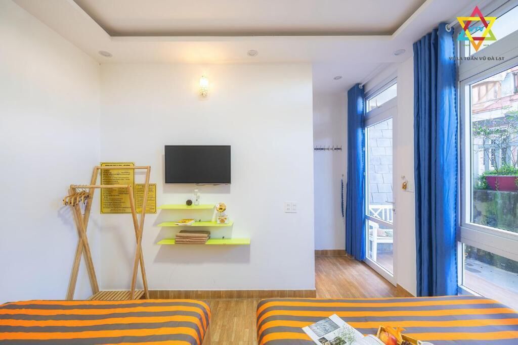 Habitación de hotel con cama, televisor montado en la pared, balcón con cortinas azules y suelo de madera.
