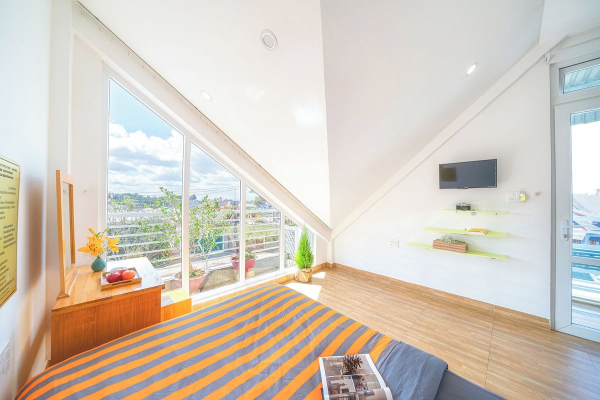 Un dormitorio moderno con un ventanal grande, una cama con ropa de cama a rayas, un televisor montado y un balcón pequeño con plantas en macetas.