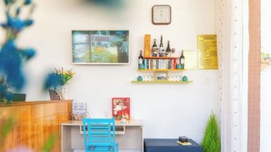 Zona de recepción con un escritorio de madera, una silla azul, un pequeño reposapiés azul, un reloj, una estantería con libros y botellas, y un cuadro enmarcado.