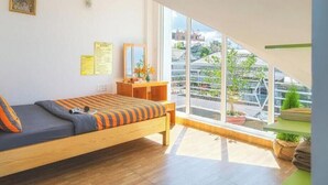 Una habitación con cama, escritorio, ventana con vista a edificios y una planta.