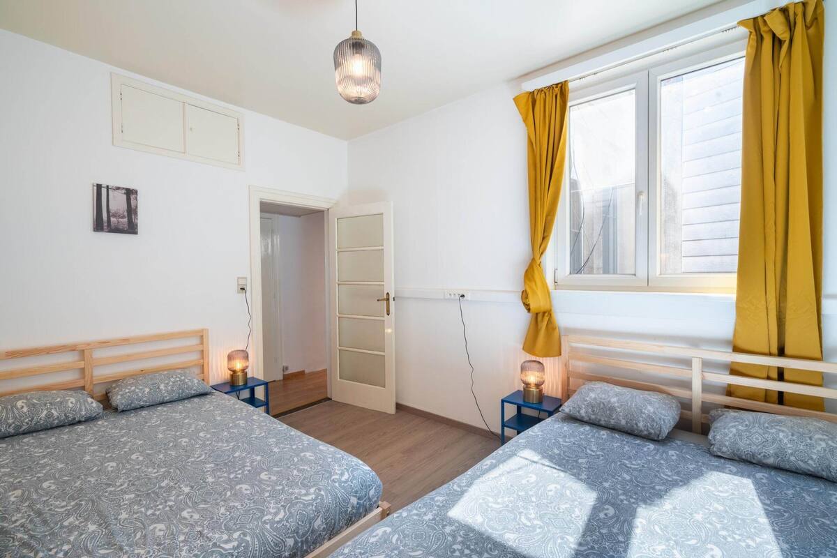 Een slaapkamer met twee bedden, een raam met gele gordijnen, een deur en een wandlamp.