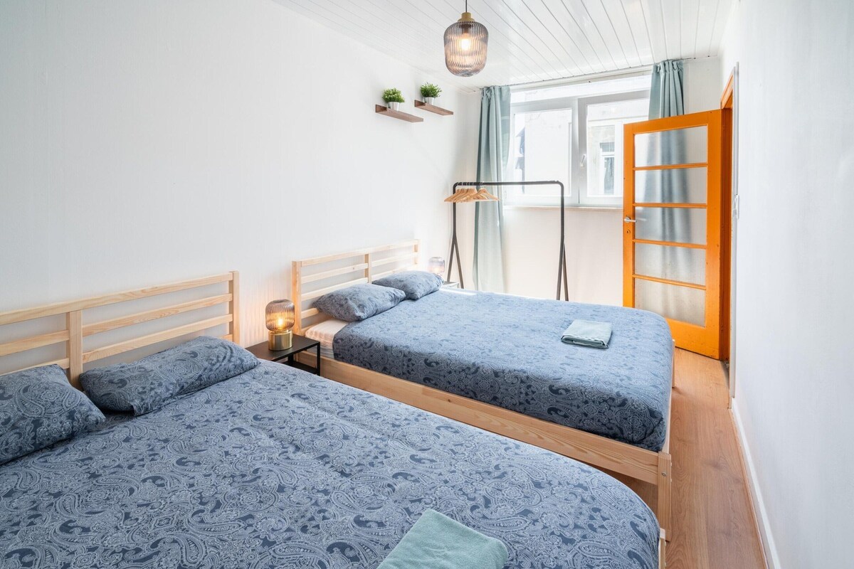 A bedroom with two beds, a nightstand, a hanging light, a window with curtains, and a wooden door.