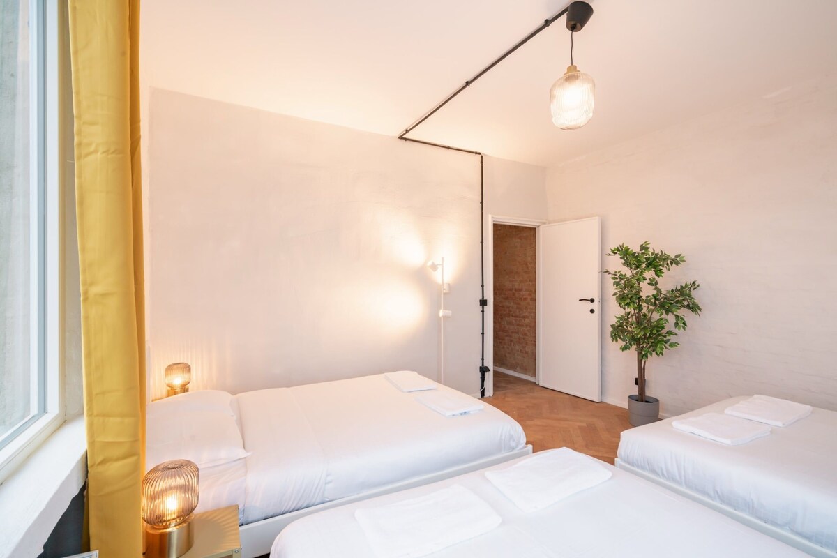Habitación de hotel con tres camas, una planta en maceta y una puerta que da a otra habitación.