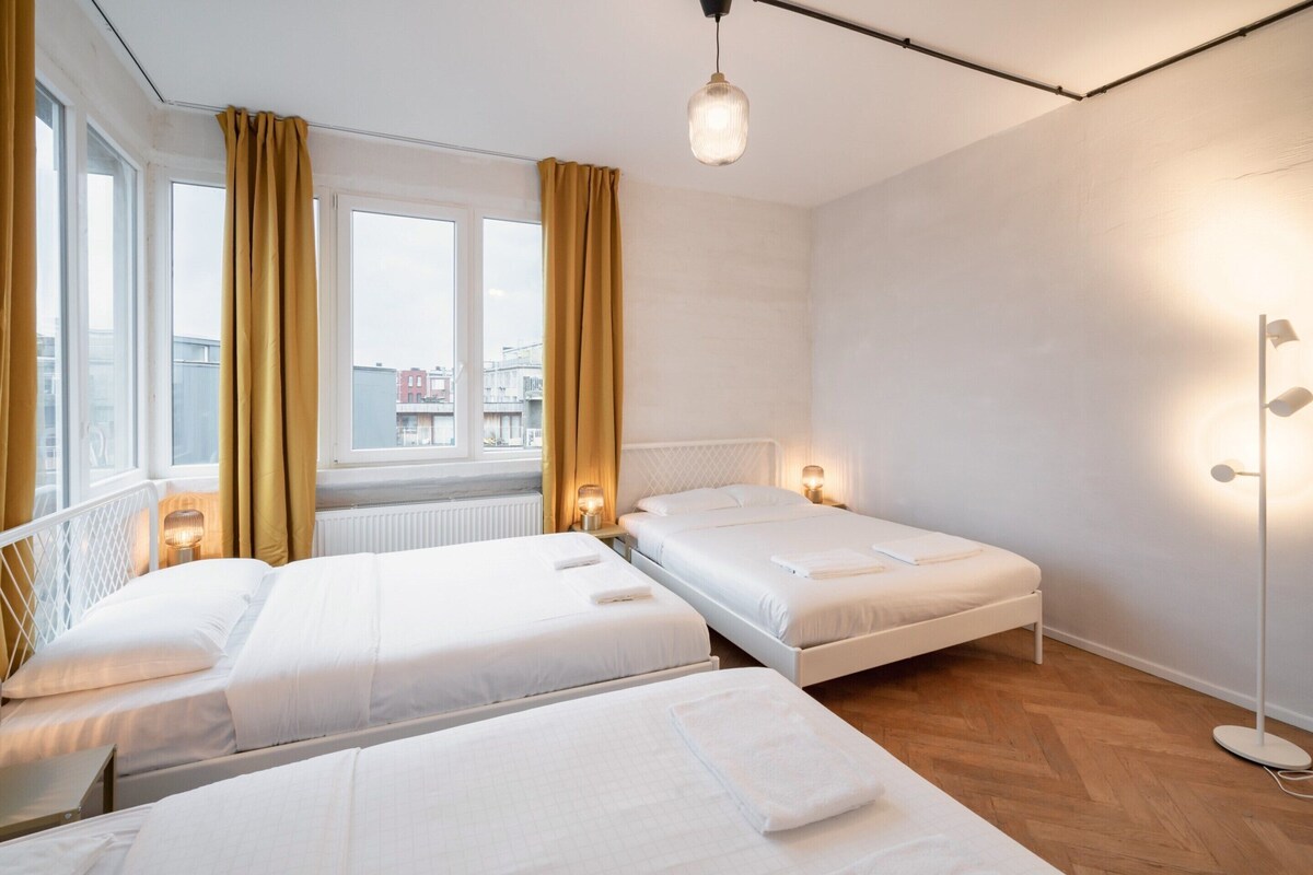 Habitación de hotel con dos camas, un ventanal con cortinas, un radiador y una lámpara de pie.