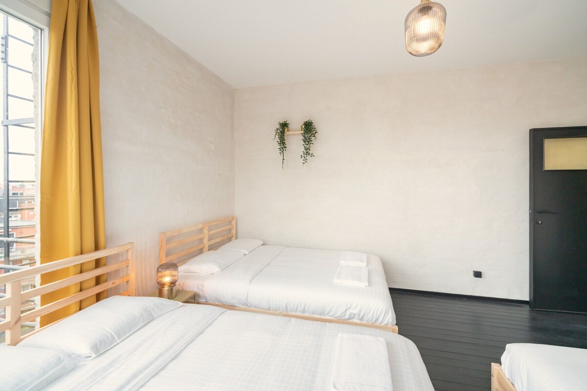 A hotel room with two beds, a wooden headboard, a hanging plant, a pendant light, and a view of buildings outside.