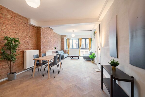 Een moderne leefruimte met een bakstenen muur, houten vloer, een zithoek met stoelen, een bank en een groot raam met gordijnen.