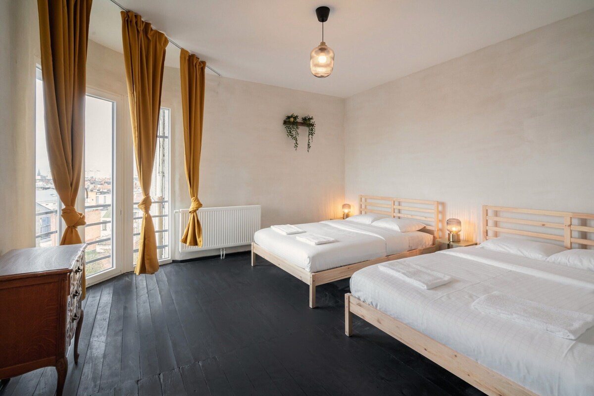A hotel room with two beds, a wooden desk, a radiator, and a view of the city through large windows.