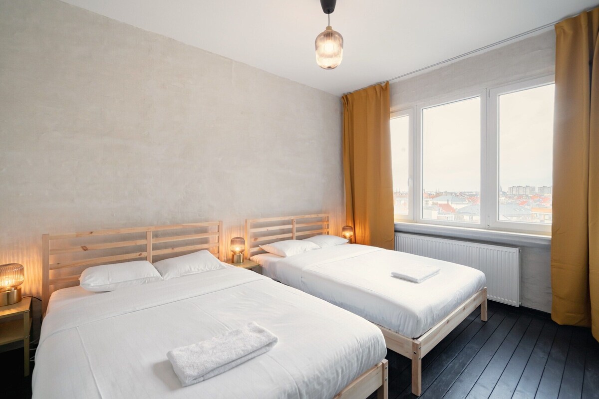 A hotel room with two beds, a large window with curtains, a radiator, and a hanging light fixture.