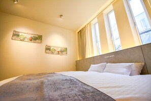 A hotel room with a bed, two wall-mounted artworks, a window with curtains, and a headboard with a light switch.