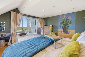 A bedroom with a large bed, a vanity with lights, a dresser, a painting, and a view of the outdoors. - Shepherds Cottage - North East Escapes (Bamburgh)
