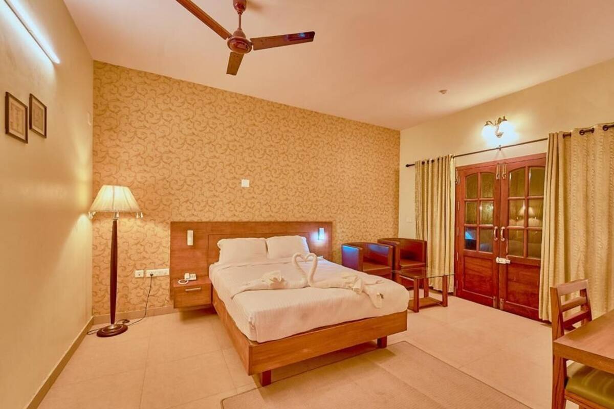 A hotel room with a bed, bedside table, lamp, ceiling fan, and a view of the outside through a door.
