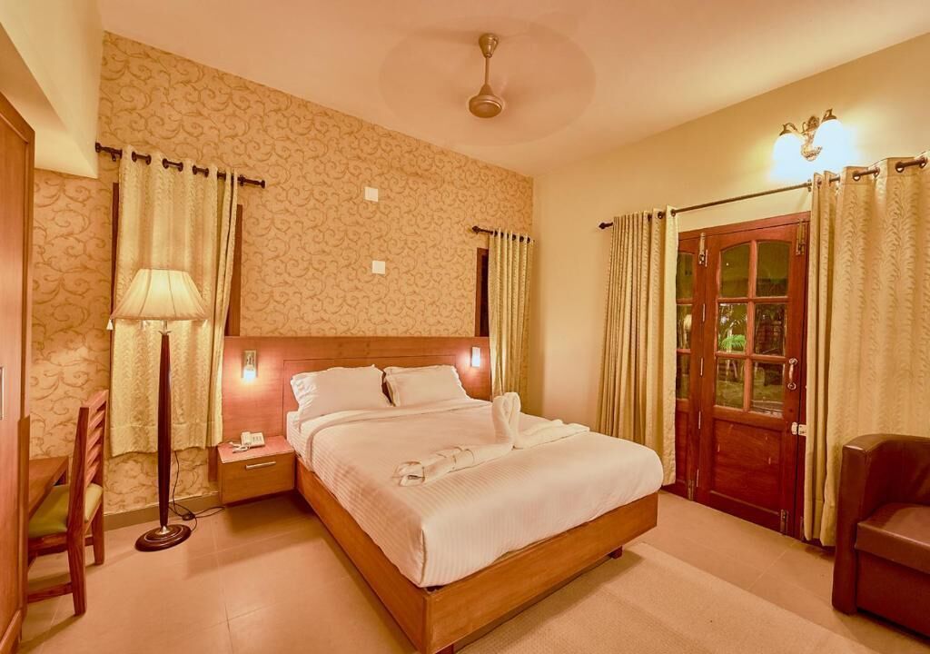 A hotel room with a bed, bedside table, lamp, and a view of the outside.