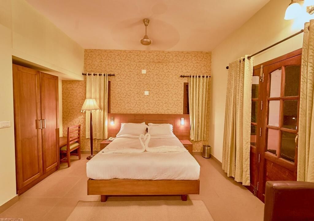 A hotel room with a large bed, wooden wardrobe, a desk with a lamp, and a window with curtains.