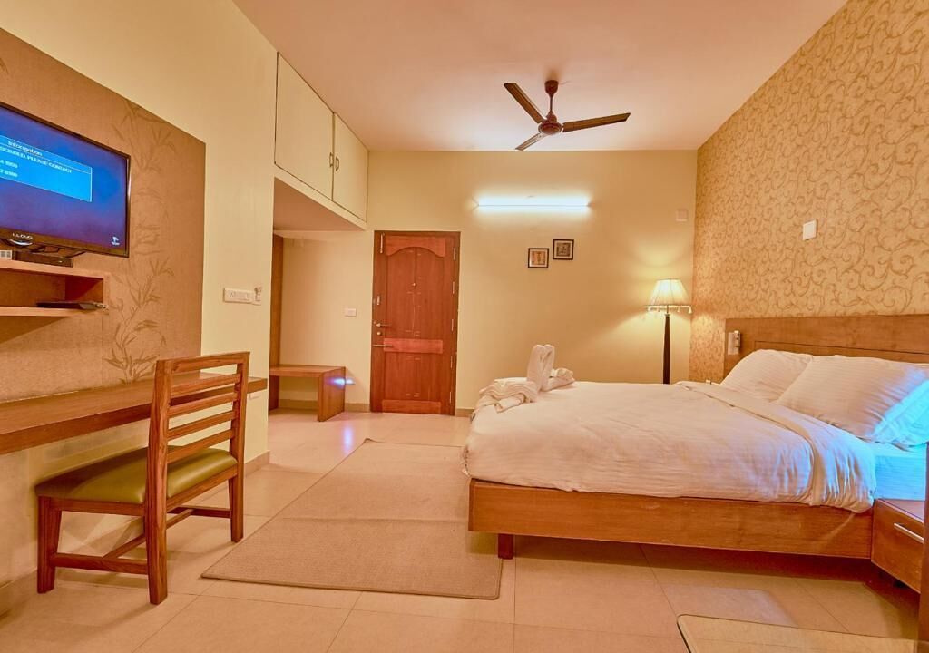 A hotel room with a bed, a desk, a chair, a television, a ceiling fan, and a door.