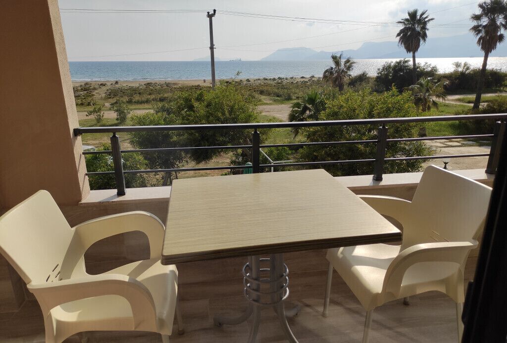 Varanda com mesa e cadeiras, vista para uma praia e palmeiras.