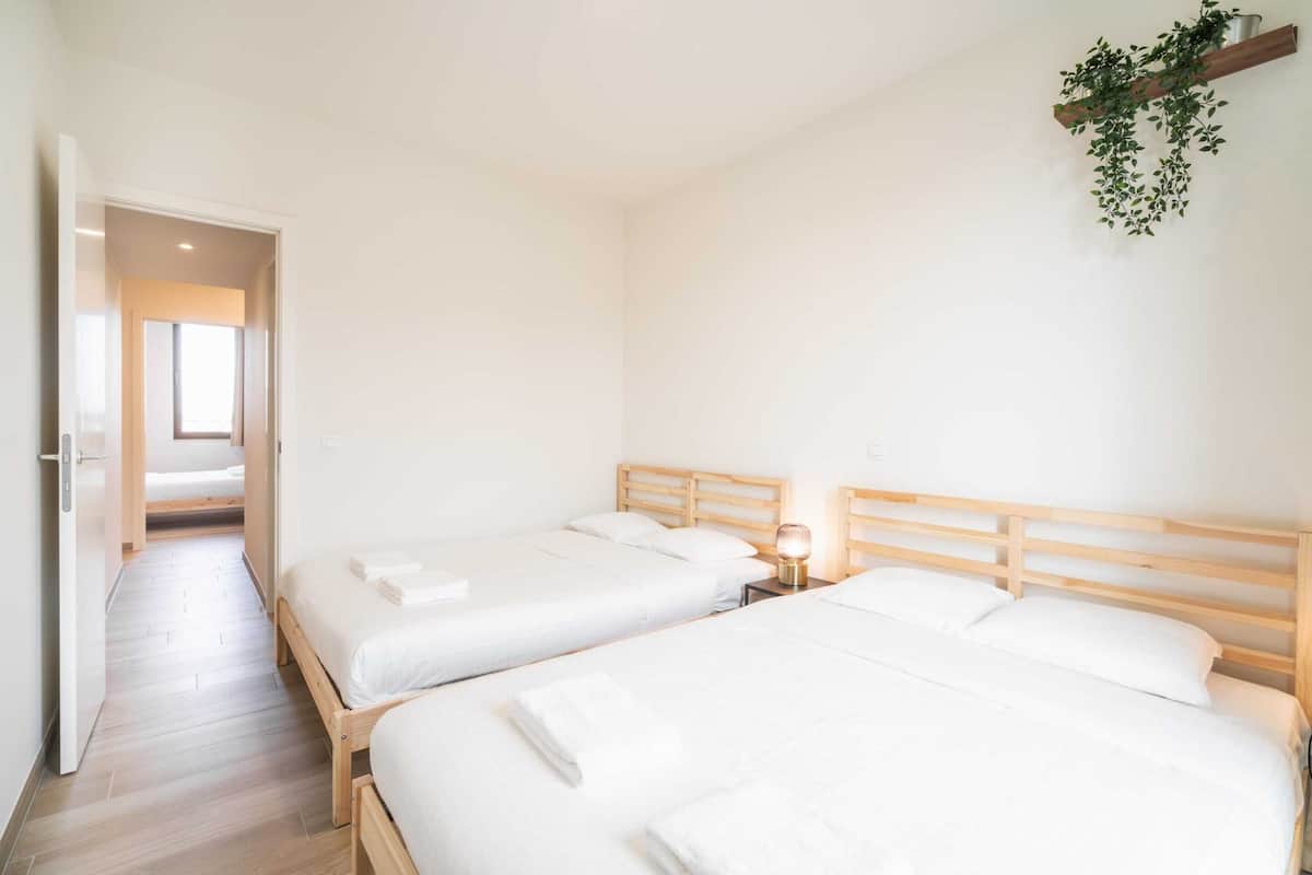 A hotel room with two beds, a wooden headboard, a bedside table with a lamp, and a hanging plant.