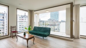 Amplia sala de estar con un sofá verde, una mesa de madera y una silla. Amplias ventanas ofrecen vistas de la ciudad.