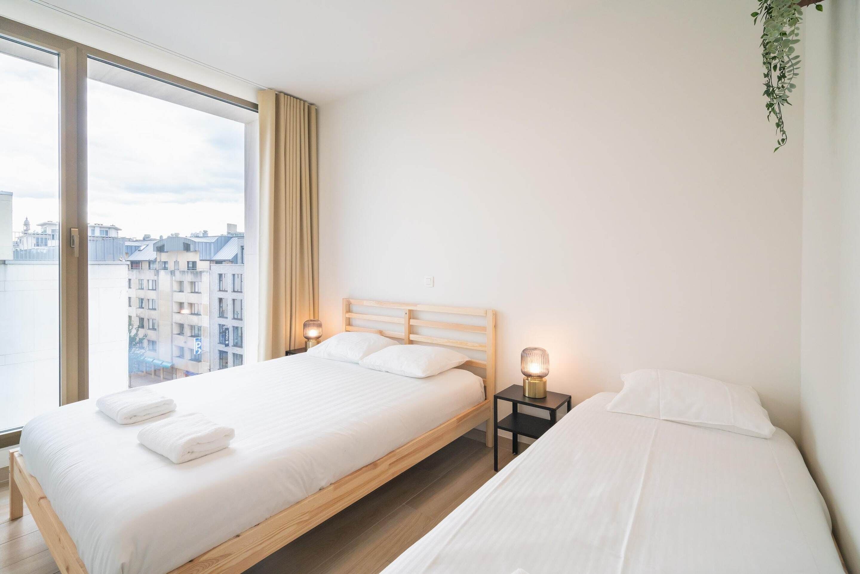 Quarto moderno com uma janela ampla, uma cama de madeira, mesa de cabeceira com abajur e vista para edifícios.