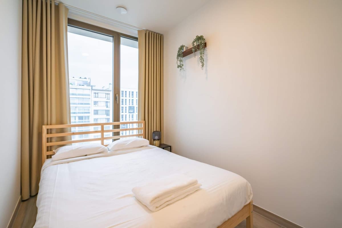A neatly made bed with a wooden headboard, a bedside table with a lamp, and a window with curtains.