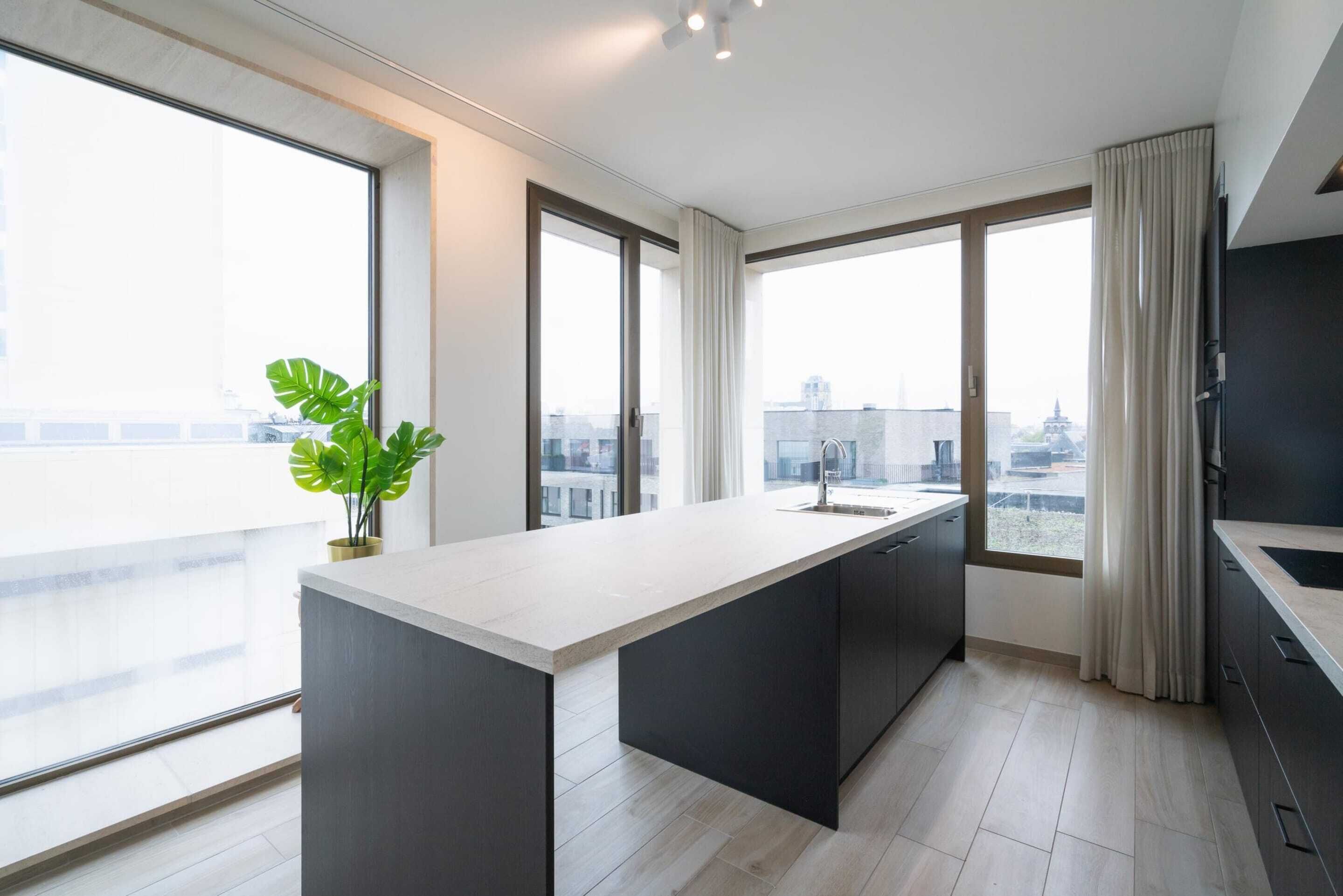A modern kitchen with a large island, dark cabinetry, and large windows offering city views.