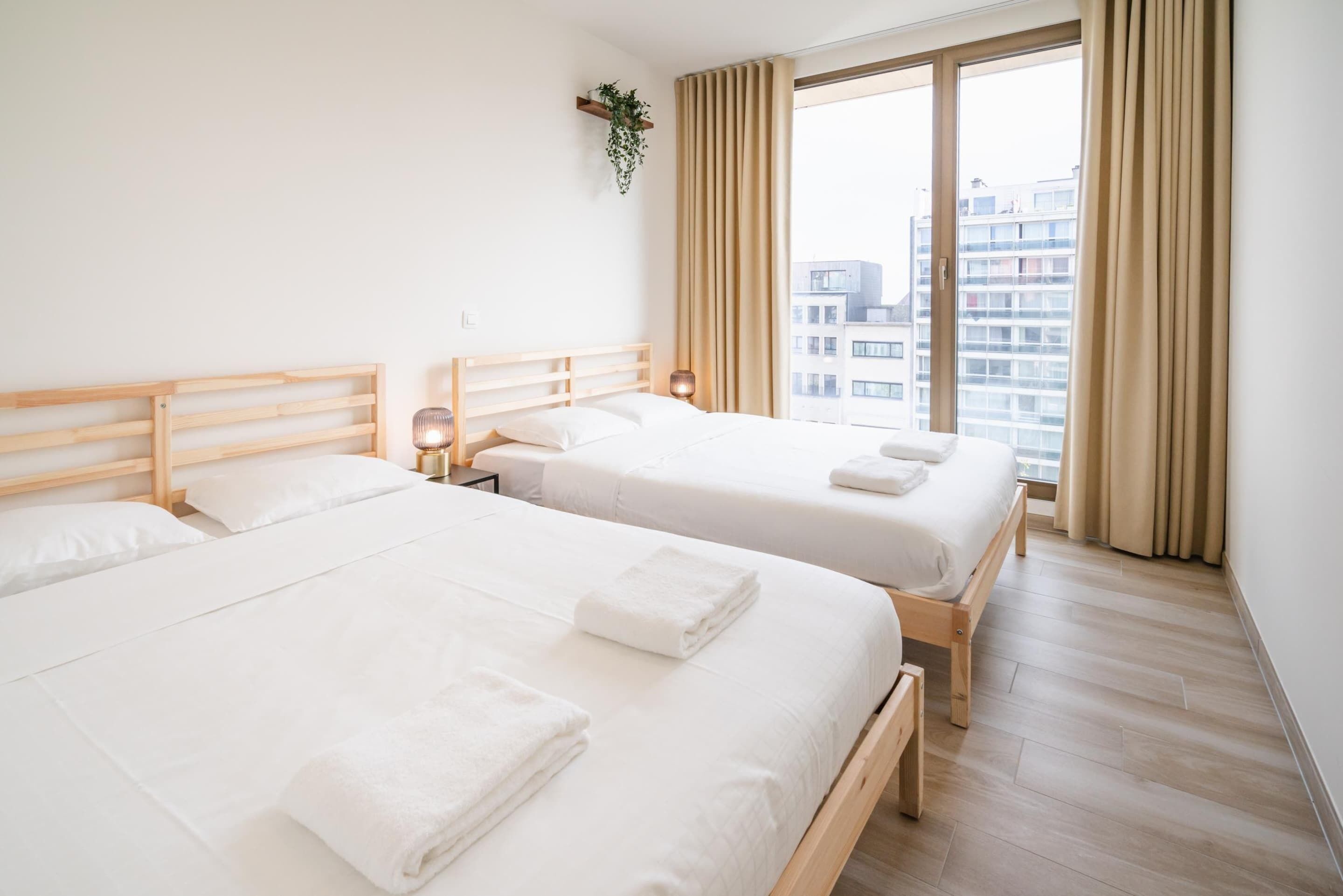 Quarto de hotel com duas camas, uma cabeceira de madeira, mesa de cabeceira com abajur e uma janela grande com cortinas.