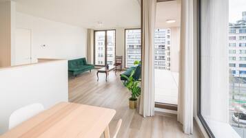 Een moderne hotelkamer met een houten eettafel, een groene bank en uitzicht op de stad door grote ramen.