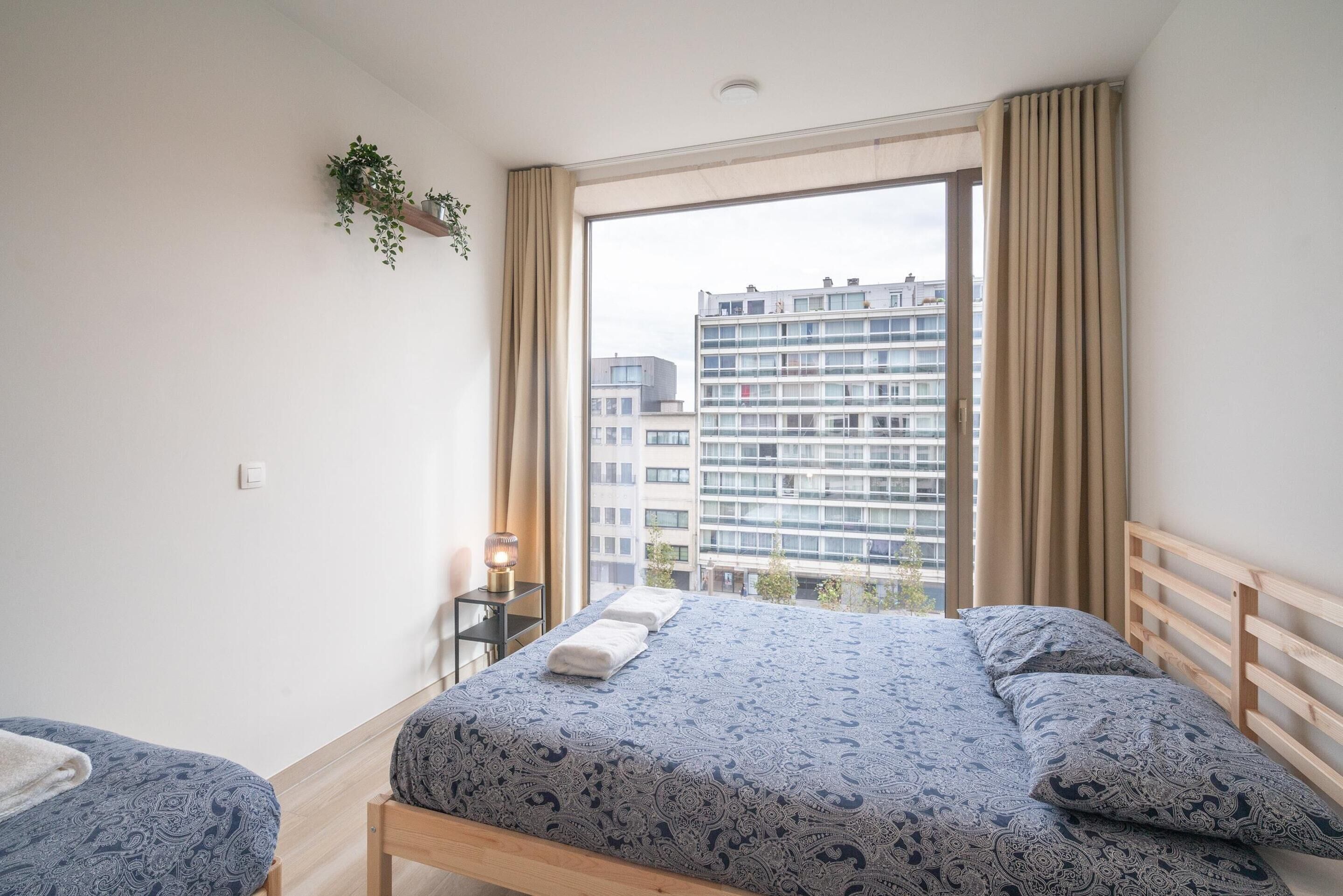 Quarto com cama, mesa de cabeceira, planta pendente e vista de edifícios pela janela.