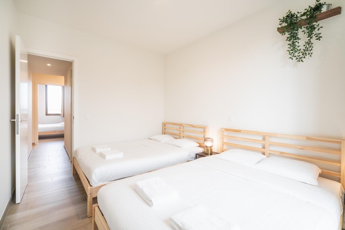 Habitación de hotel con dos camas, cabecera de madera, mesita de noche con lámpara y una planta colgante.