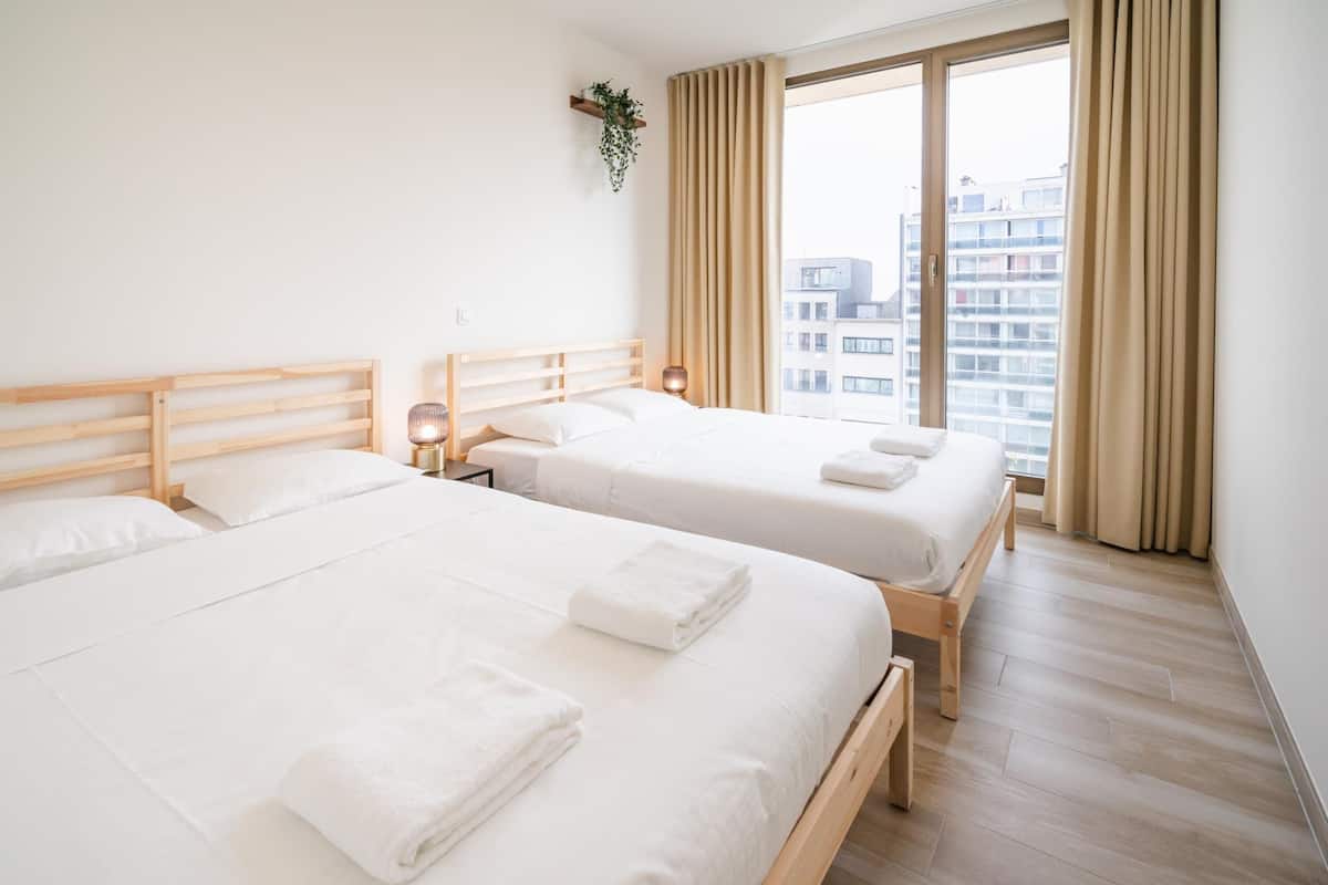 A hotel room with two beds, a wooden headboard, a bedside table with a lamp, and a large window with curtains.