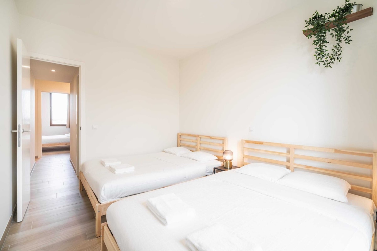 A hotel room with two beds, a wooden headboard, a bedside table with a lamp, and a hanging plant.