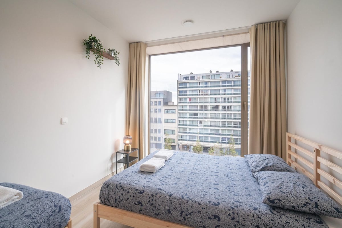 A bedroom with a bed, a bedside table, a hanging plant, and a view of buildings through a window.