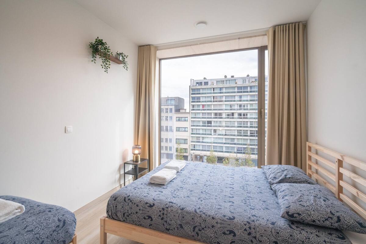A bedroom with a bed, a bedside table, a hanging plant, and a view of buildings through a window.