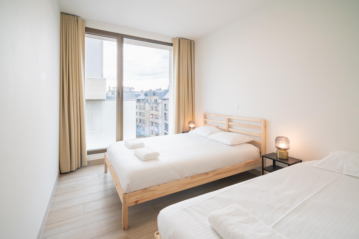 A hotel room with a large window, a wooden bed, a bedside table with a lamp, and a second bed.