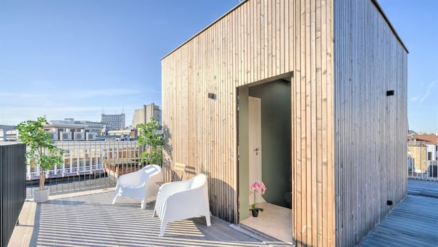 A rooftop terrace with a wooden structure, two white chairs, a potted plant, and a view of the cityscape.