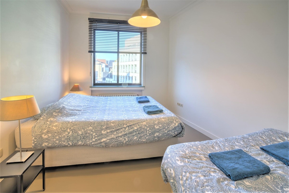 A bedroom with a bed, a bedside table, a lamp, a window with blinds, and a view of buildings outside.